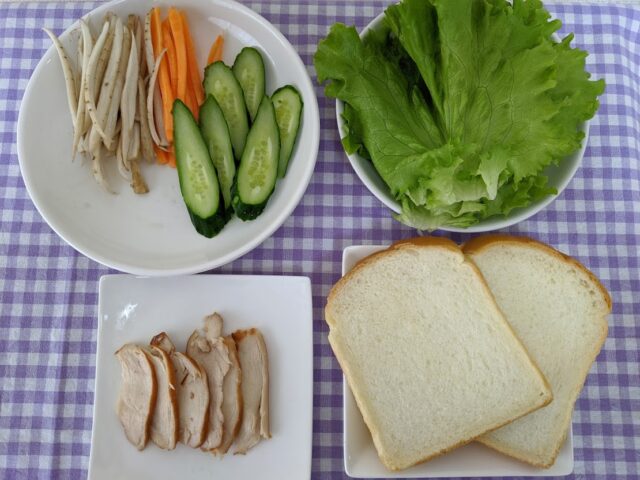 ごぼうサラダとチキンのサンドイッチの材料でごぼうとにんじんとレタスとチキンと食パンがテーブルの上に並んでいる写真