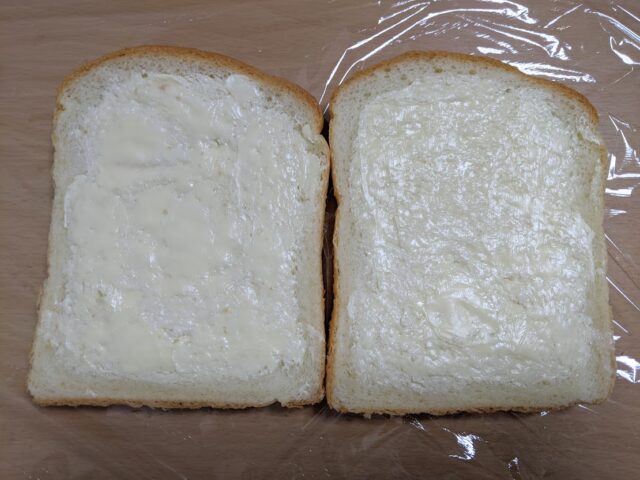 ラップを敷いたテーブルの上にバターをぬった食パン2枚を置いている写真
