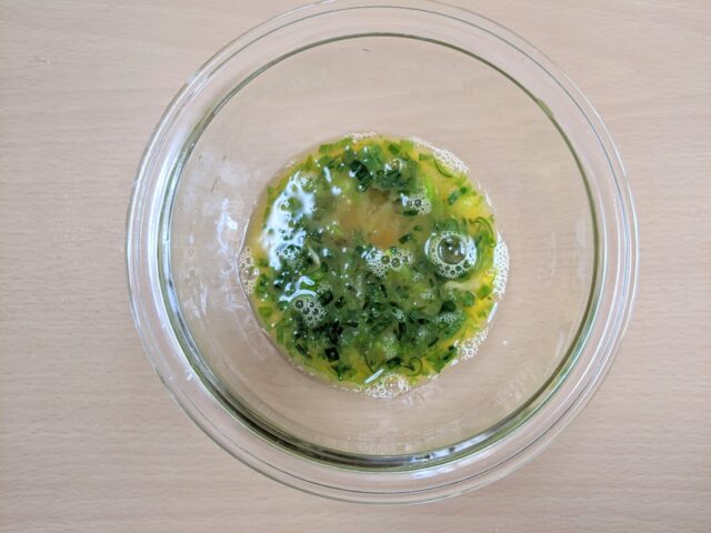 ガラスボウルに溶き卵とたまねぎの葉のみじん切りと調味料を加え混ぜたものをテーブルに置いている写真