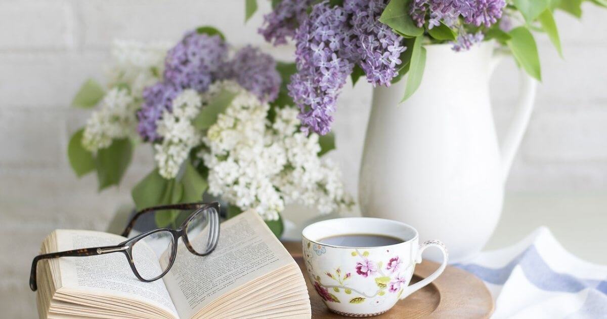 テーブルに置いてあるコーヒーが入ったコーヒーカップと本と眼鏡と紫と白のお花が花瓶に生けられている画像