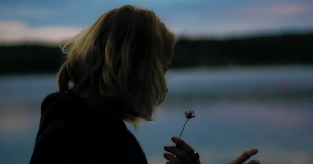 薄暗い空の背景で女性が川辺で一輪の花を手にもって下を向いている画像