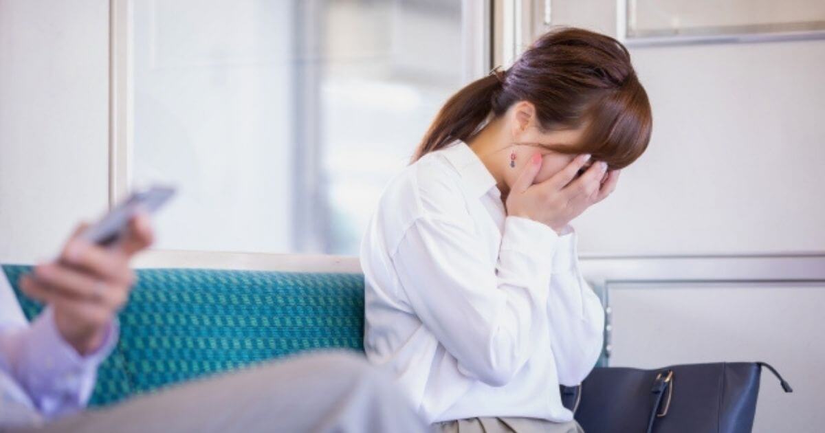 女性が電車の中で両手を顔に当てて疲れている画像