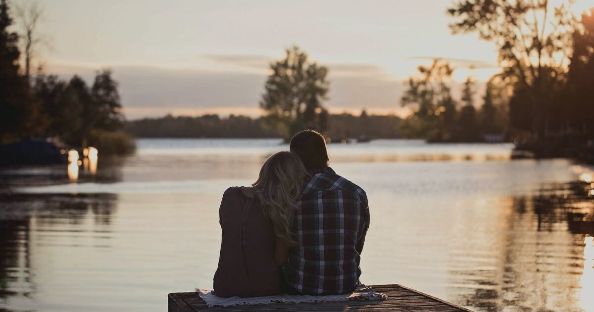 川沿いにカップルで座り夕日を眺めている画像